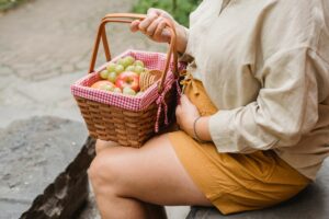 Crop anonymous female in expectancy wearing casual clothes with basket full of ripe fruits and biscuits prepared for picnic